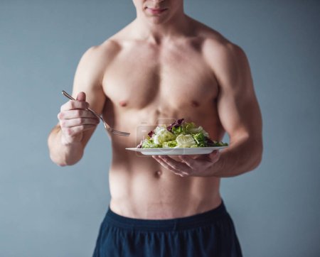 Téléchargez les photos : Image recadrée d'un beau mec musclé avec un torse nu mangeant de la salade, sur fond gris - en image libre de droit