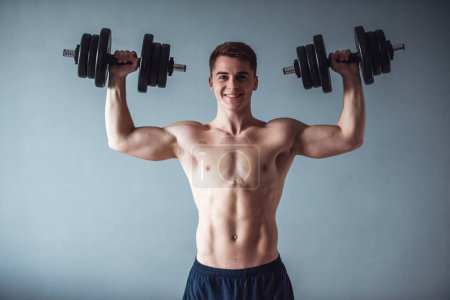 Foto de Tipo musculoso guapo con el torso desnudo está sosteniendo pesas, mirando a la cámara y sonriendo, sobre fondo gris - Imagen libre de derechos