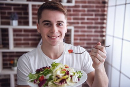 Téléchargez les photos : Beau jeune homme regarde la caméra et sourit tout en mangeant de la salade dans la cuisine à la maison - en image libre de droit