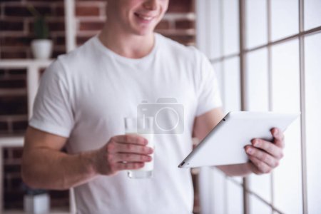 Téléchargez les photos : Beau jeune homme boit du lait, en utilisant une tablette numérique et souriant tout en restant debout dans la cuisine à la maison - en image libre de droit