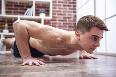 Foto de Guapo musculoso chico con torso desnudo está haciendo flexiones mientras hace ejercicio en casa - Imagen libre de derechos