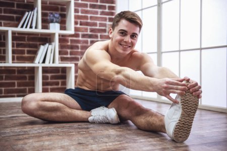 Foto de Tipo musculoso guapo con el torso desnudo se estira, mirando a la cámara y sonriendo mientras se entrena en casa - Imagen libre de derechos