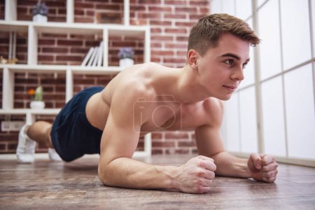 Foto de Chico musculoso guapo con torso desnudo está haciendo ejercicio de tablón mientras hace ejercicio en casa - Imagen libre de derechos
