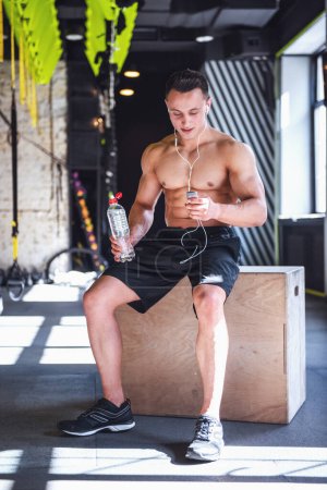 Téléchargez les photos : Beau jeune sportif musclé écoute de la musique, tenant une bouteille d'eau et souriant après l'entraînement dans la salle de gym - en image libre de droit