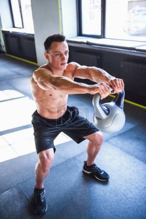 Photo for Handsome young muscular sportsman is working out with kettlebell in gym - Royalty Free Image