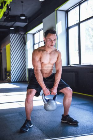 Photo for Handsome young muscular sportsman is working out with kettlebell in gym - Royalty Free Image