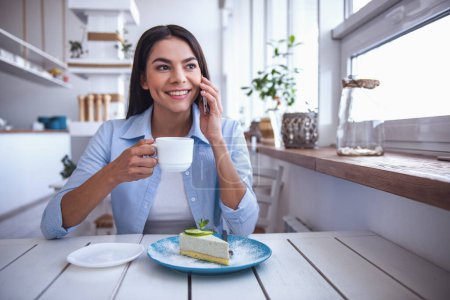 Foto de La joven hermosa chica bebiendo café y postre comiendo, sentada en un café en la terraza y hablando por teléfono - Imagen libre de derechos