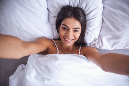 Téléchargez les photos : Belle fille gaie au lit fait selfie - en image libre de droit