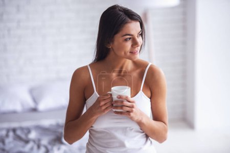 Foto de ¡Buenos días! Feliz joven hermosa mujer bebiendo té o café en casa en el dormitorio
. - Imagen libre de derechos