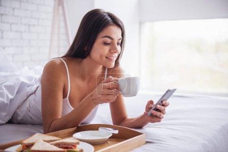 Photo for Good morning! Happy young beautiful woman eating breakfast while sitting on the bed at home - Royalty Free Image