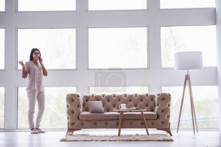 Foto de Oficina central. Exitosa joven hermosa mujer de pie cerca de una gran ventana en la sala de estar y hablando por teléfono - Imagen libre de derechos