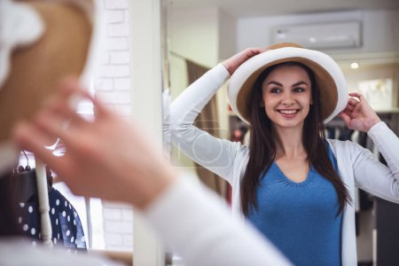 Foto de Hermosa chica está eligiendo un sombrero, mirando en el espejo y sonriendo mientras hace compras en la boutique - Imagen libre de derechos
