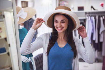 Foto de Hermosa chica está eligiendo un sombrero y sonriendo mientras hace compras en boutique - Imagen libre de derechos