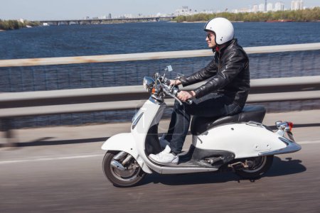 Photo for Guy in leather jacket and helmet is riding on scooter through the city bridge and smiling - Royalty Free Image