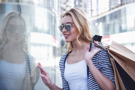 Foto de Hermosa chica en gafas de sol está sosteniendo bolsas de compras, mirando escaparates y sonriendo mientras camina por la calle - Imagen libre de derechos