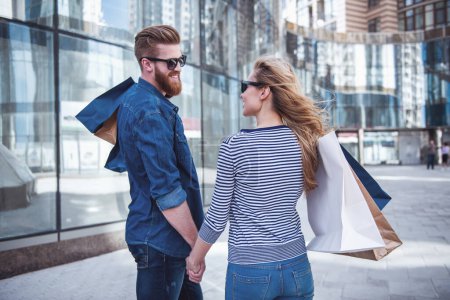 Photo for Beautiful young couple in sun glasses and with shopping bags holding hands, looking at each other and smiling while walking down the street - Royalty Free Image