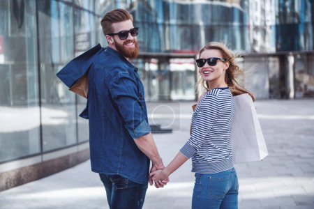 Téléchargez les photos : Beau jeune couple dans des lunettes de soleil et avec des sacs à provisions tenant la main, regardant la caméra et souriant tout en marchant dans la rue - en image libre de droit
