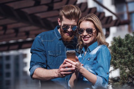 Téléchargez les photos : Beau jeune couple en lunettes de soleil utilise un téléphone intelligent et souriant tout en se tenant debout à l'extérieur - en image libre de droit