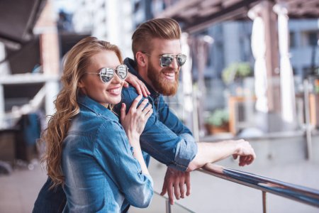 Téléchargez les photos : Beau jeune couple dans des lunettes de soleil est étreignant, regardant loin et souriant tout en se tenant sur le balcon - en image libre de droit