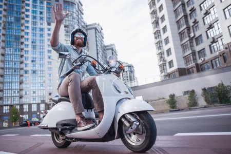 Téléchargez les photos : Beau barbu guy en lunettes de soleil et casque est agitant et souriant tout en conduisant un scooter - en image libre de droit