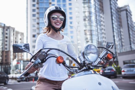 Téléchargez les photos : Belle fille dans les lunettes de soleil et le casque est souriant tout en montant un scooter - en image libre de droit