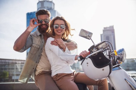Téléchargez les photos : Beau jeune couple en lunettes de soleil regarde la caméra et souriant tout en étant assis sur un scooter à l'extérieur - en image libre de droit