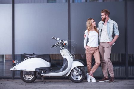 Téléchargez les photos : Beau jeune couple se serre dans ses bras, se regardant et souriant tout en se tenant près du scooter à l'extérieur - en image libre de droit
