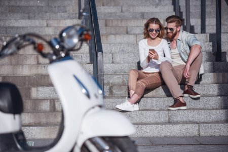 Téléchargez les photos : Beau jeune couple en lunettes de soleil utilise un téléphone intelligent et souriant tout en étant assis sur les escaliers à l'extérieur - en image libre de droit