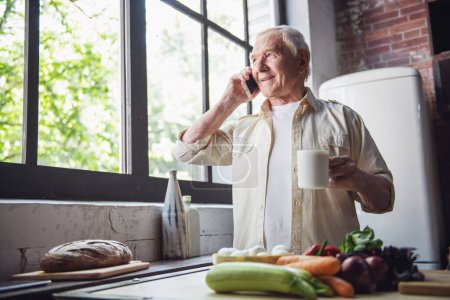 Téléchargez les photos : Beau vieillard tient une tasse, parle sur le téléphone portable et sourit tout en se tenant dans la cuisine - en image libre de droit