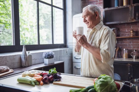 Téléchargez les photos : Beau vieillard tient une tasse, sentant l'arôme et souriant tout en se tenant dans la cuisine - en image libre de droit