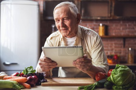 Foto de Hombre mayor guapo en delantal está utilizando una tableta digital y sonriendo mientras cocina en la cocina - Imagen libre de derechos