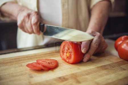 Téléchargez les photos : Image recadrée d'un bel homme âgé coupant des tomates pendant la cuisson dans la cuisine - en image libre de droit