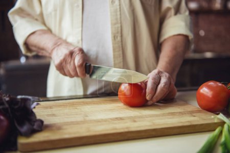 Téléchargez les photos : Image recadrée d'un bel homme âgé coupant des tomates pendant la cuisson dans la cuisine - en image libre de droit