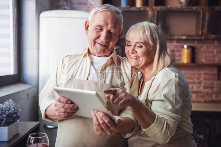 Foto de Hermosa pareja de ancianos en delantales está utilizando una tableta digital, hablando y sonriendo mientras cocinan juntos en la cocina - Imagen libre de derechos