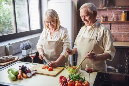 Photo for Beautiful senior couple in aprons is talking and smiling while cooking together in kitchen - Royalty Free Image