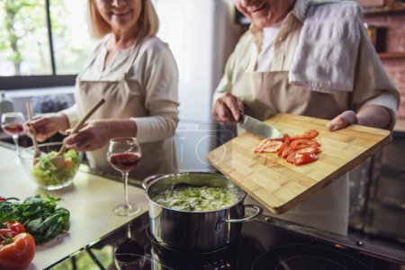 Téléchargez les photos : Beau couple de personnes âgées dans des tabliers parle et sourit tout en cuisinant ensemble dans la cuisine - en image libre de droit