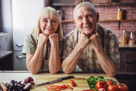 Photo for Beautiful senior couple in aprons is talking and smiling while cooking together in kitchen - Royalty Free Image