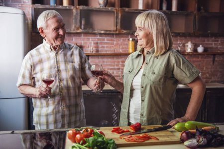 Téléchargez les photos : Beau couple de personnes âgées boit du vin et sourit tout en cuisinant ensemble dans la cuisine - en image libre de droit