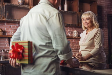 Foto de Mujer mayor hermosa está cocinando y sonriendo mientras su marido está sosteniendo un regalo para ella a sus espaldas - Imagen libre de derechos