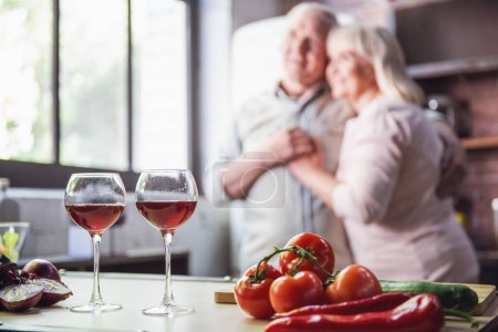 Foto de Hermosa pareja de ancianos está bailando y sonriendo mientras cocinan juntos en la cocina. Vasos de vino en primer plano - Imagen libre de derechos