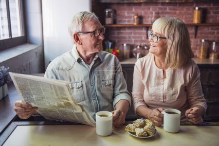 Téléchargez les photos : Beau couple âgé boit du thé, lit le journal et sourit tout en se reposant dans la cuisine - en image libre de droit