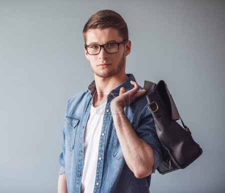 Photo for Handsome young man in casual clothes and glasses is holding a stylish bag and looking at camera, on gray background - Royalty Free Image