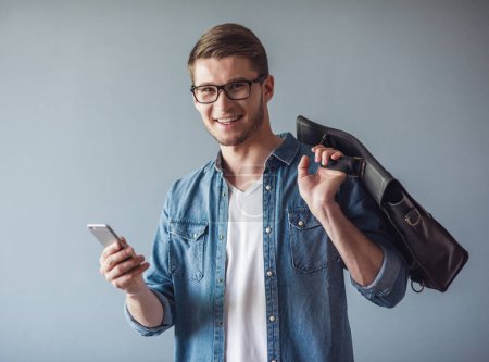 Téléchargez les photos : Beau jeune homme d'affaires en vêtements décontractés, lunettes et avec un sac élégant utilise un téléphone intelligent et souriant, sur fond gris - en image libre de droit