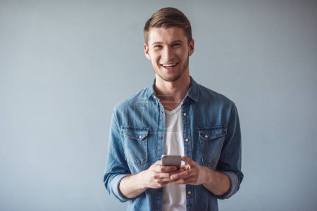 Téléchargez les photos : Beau gars en vêtements décontractés utilise un téléphone intelligent, regardant la caméra et souriant, sur fond gris - en image libre de droit