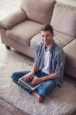 Photo for Handsome guy is using a laptop, looking at camera and smiling while sitting on the floor at home - Royalty Free Image