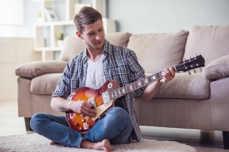 Téléchargez les photos : Beau gars joue de la guitare et sourit tout en étant assis sur le sol à la maison - en image libre de droit