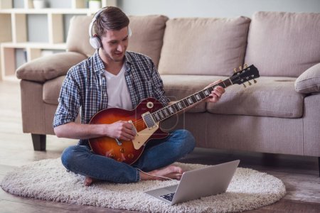 Foto de Chico guapo en auriculares está tocando la guitarra eléctrica, usando un ordenador portátil y sonriendo mientras está sentado en el suelo en casa - Imagen libre de derechos