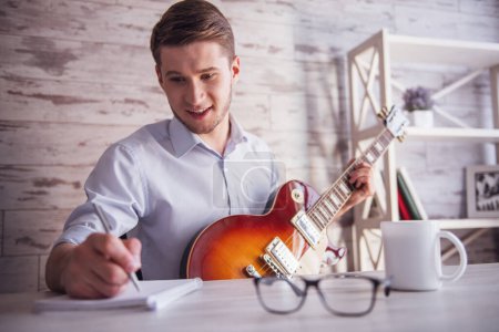 Téléchargez les photos : Beau jeune homme d'affaires joue de la guitare, prend des notes et sourit dans son bureau pendant la pause - en image libre de droit