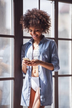 Téléchargez les photos : Belle fille en lingerie blanche et chemise en jean déboutonnée utilise un téléphone intelligent et souriant tout en se tenant près de la fenêtre - en image libre de droit