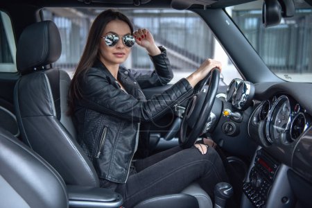 Photo for Beautiful girl in leather jacket and sun glasses is looking at camera while driving a car - Royalty Free Image